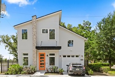 Two‑story modern house with a light exterior, tall pitched rooflines, a vertical stone accent column, and an orange front door. A small driveway with a parked car is on the right side, and the house is surrounded by trees and shrubs under a bright blue sky.